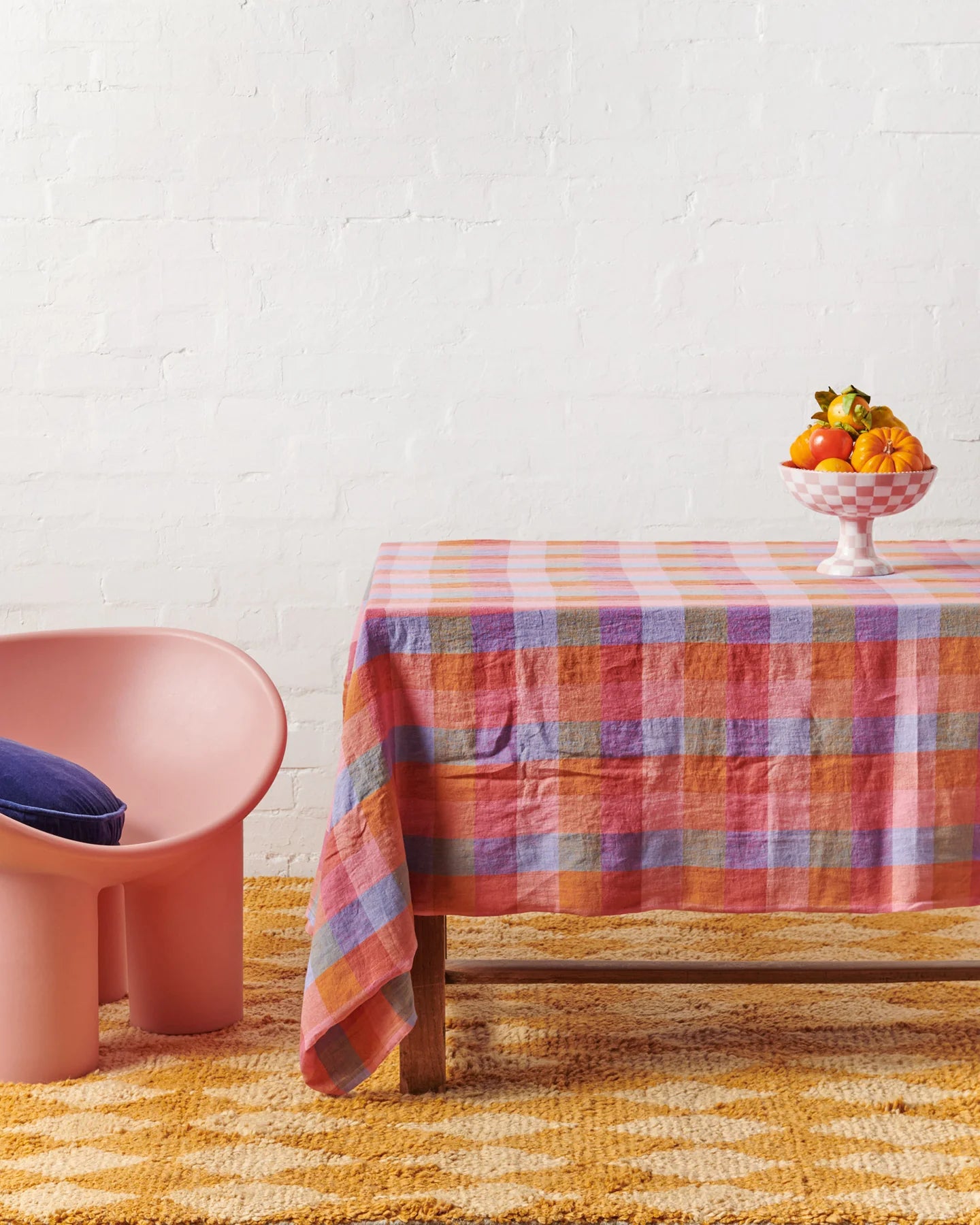 Tartan pattern tablecloth laid out on timber table with ceramic bowl on top filled with assorted vegetables. Sat next to table is pink chair and mustard geometric rug on floor.