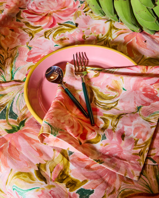 A styled flat-lay of a pink ceramic plate with a yellow rim, set on a pink floral tablecloth with a matching napkin, gold-toned cutlery, and green leaves.