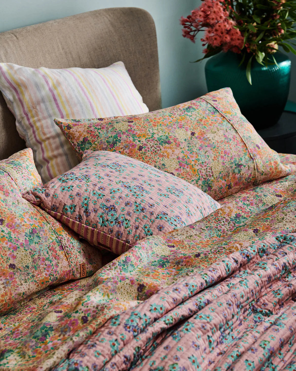 Floral patterned bedding and pillows on a bed with a vase of flowers in the background.