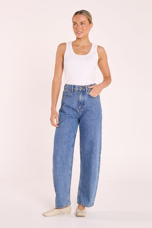 A woman modeling a pair of high-waisted, wide-leg blue jeans with a white tank top and beige shoes, standing against a solid light beige studio background.