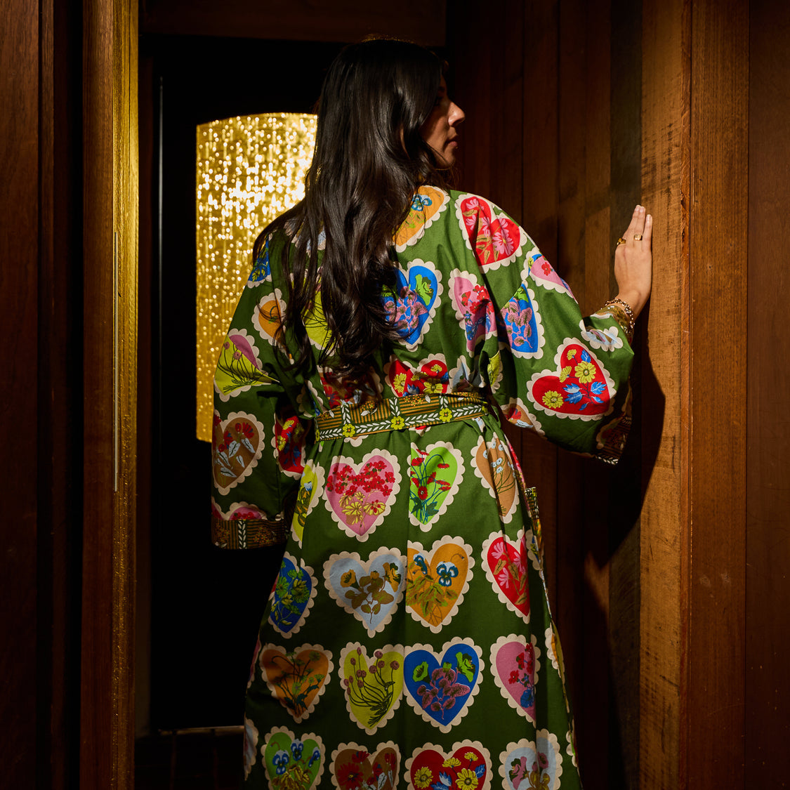 A woman viewed from behind wearing a green kimono-style robe with a colorful heart pattern, standing in a dark wooden doorway.