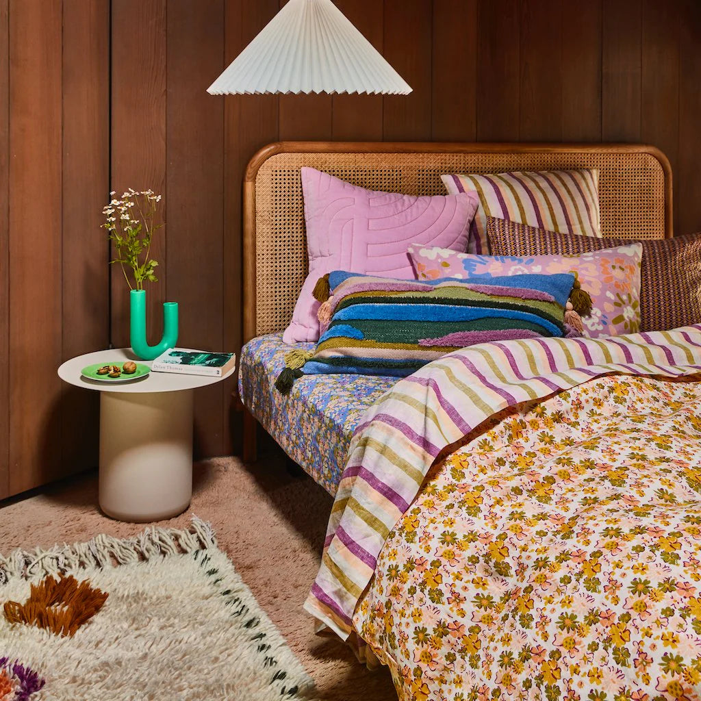 Timber wall, white pendant light, rattan timber bed head, white side table with green u shape vase and flower sprigs inside. Bed is covered with Loveat bed sheet and quilt cover, assorted colourful cushions.
