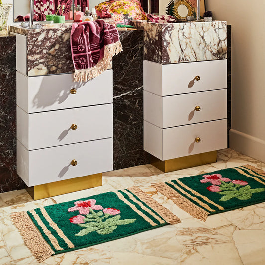 Two white bathroom vanities with marble top and gold accents, accompanied by floral-patterned bath mats on a tiled floor. Bath mat colours are green base with lighter green leaves and pink flowers, cream stripes and cream fringing along sides of bath mats.