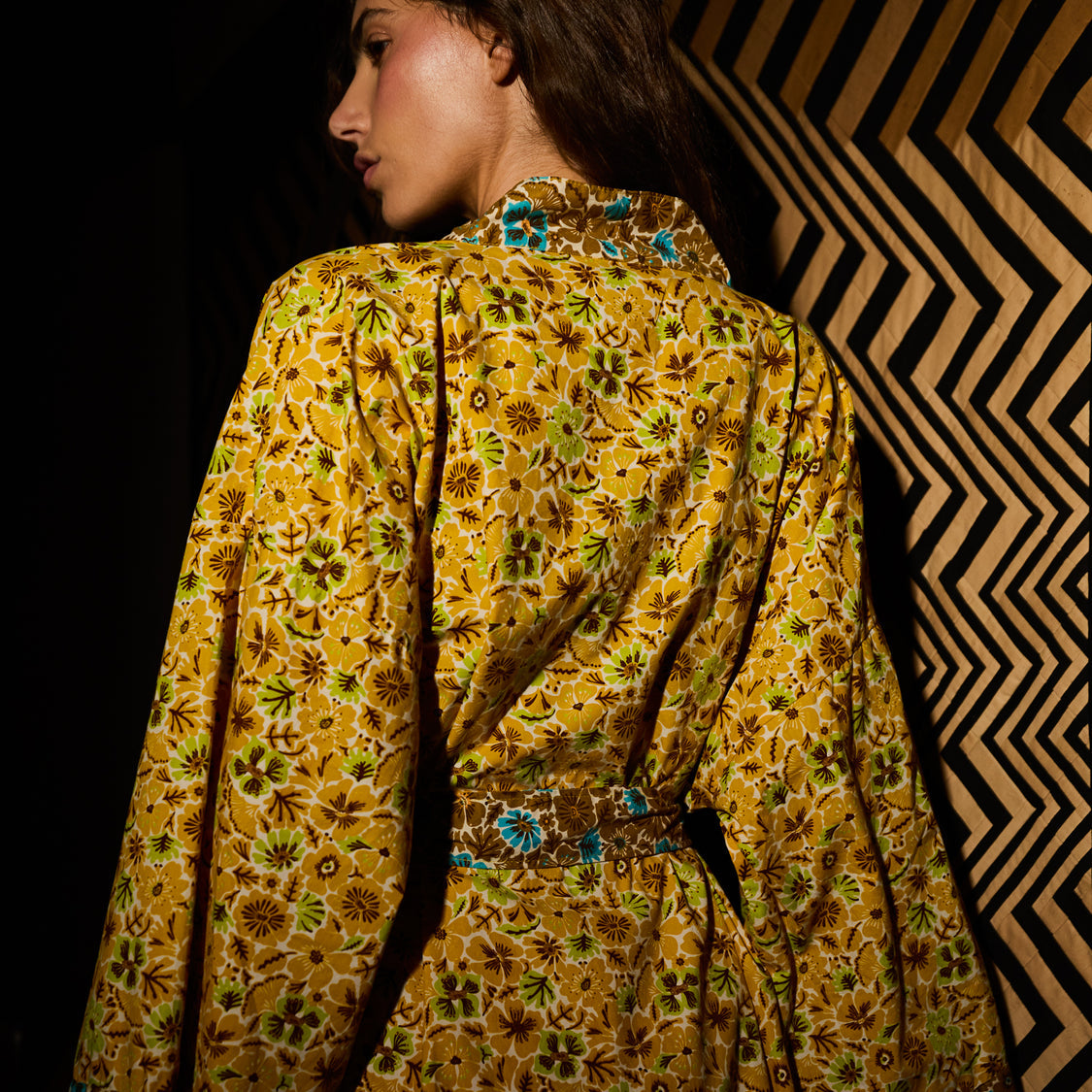 A woman shown from the back wearing a mustard yellow floral robe with green and brown patterns and a matching belt, set against a dark background with a geometric patterned screen.