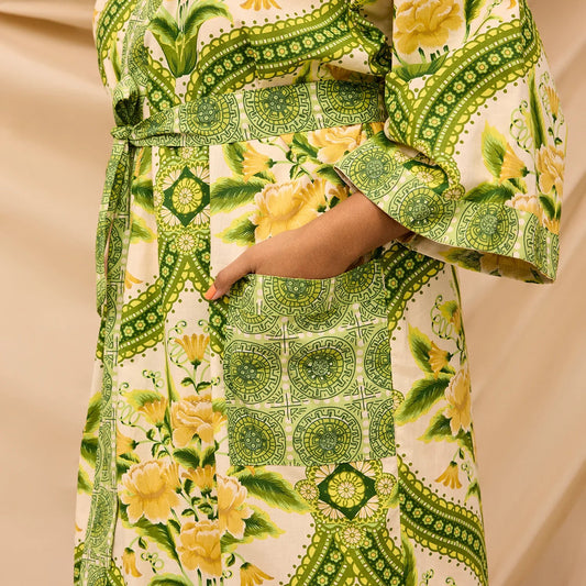 A close-up of a person wearing a patterned kimono-style robe or dress with wide sleeves and a waist tie. The fabric has a cream base with green geometric borders and yellow floral motifs, and the person's hand is visible in a side pocket.