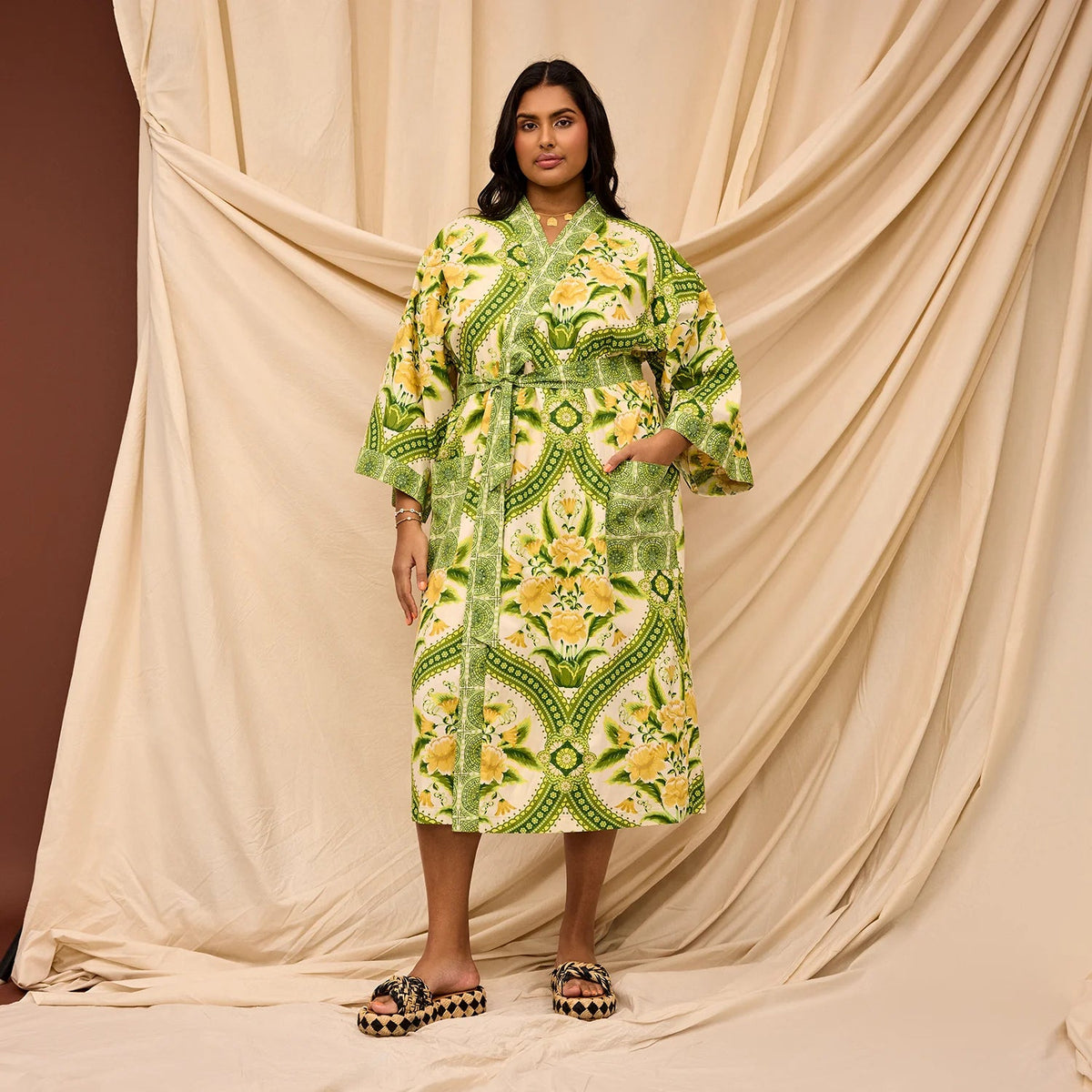 A woman stands in front of draped beige fabric wearing a long-sleeve kimono-style robe with a green, yellow, and white geometric floral pattern, paired with patterned sandals.