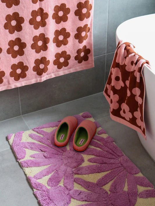 Bathroom with pink and brown floral towels on a gray tiled wall, purple bath mat with floral pattern on floor, and red slippers.