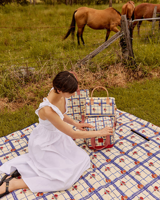 Woman in a white dress sitting on a checkered blanket with a plaid handbag, surrounded by horses in a field.