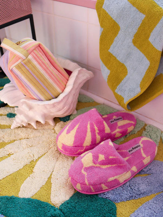 Pink and yellow patterned slippers on a colorful rug with a shell and striped towel in the background.