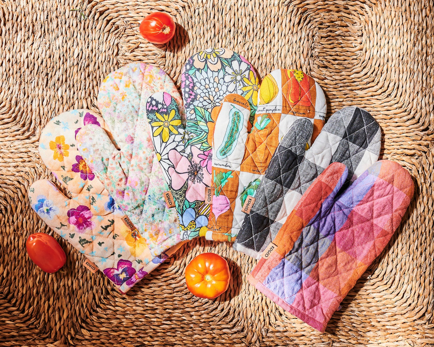 Multiple oven mitts in different colours and prints laid out on natural woven mat and scattered tomatoes