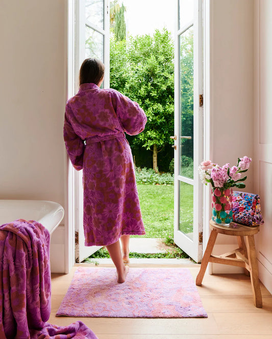 Person wearing a purple robe standing in a room with a large open door leading to a garden. Shot features Peach Fleur bath mat on floor.