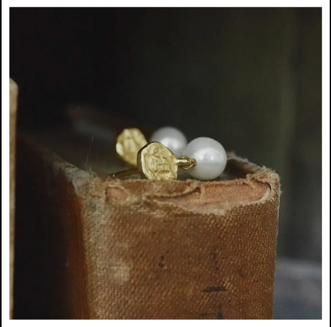 Gold circle stud and pearl earring on a textured brown surface with a dark background.