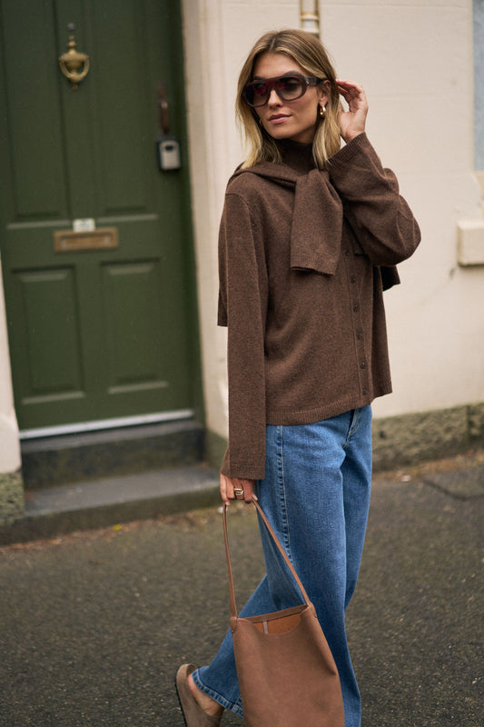 Woman wearing a brown sweater and blue jeans, holding a brown bag, standing in front of a green door.