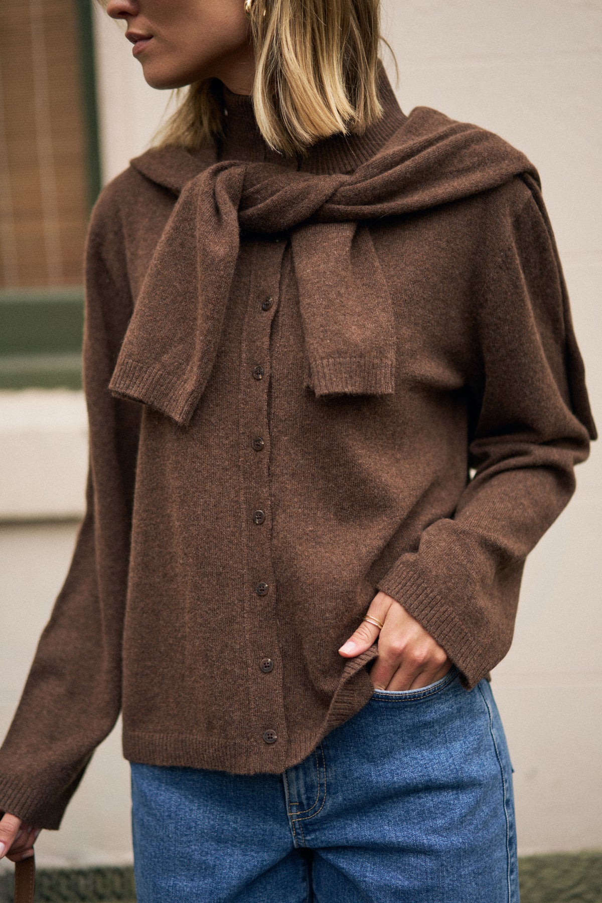 Person wearing a brown sweater and scarf with blue jeans indoors.