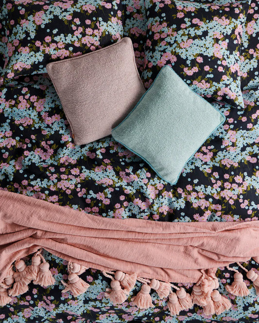 Floral patterned fabric with pink tassel throw and two cushions on top. Bedding pattern is black base with blue and pink dainty flowers with green leaf.