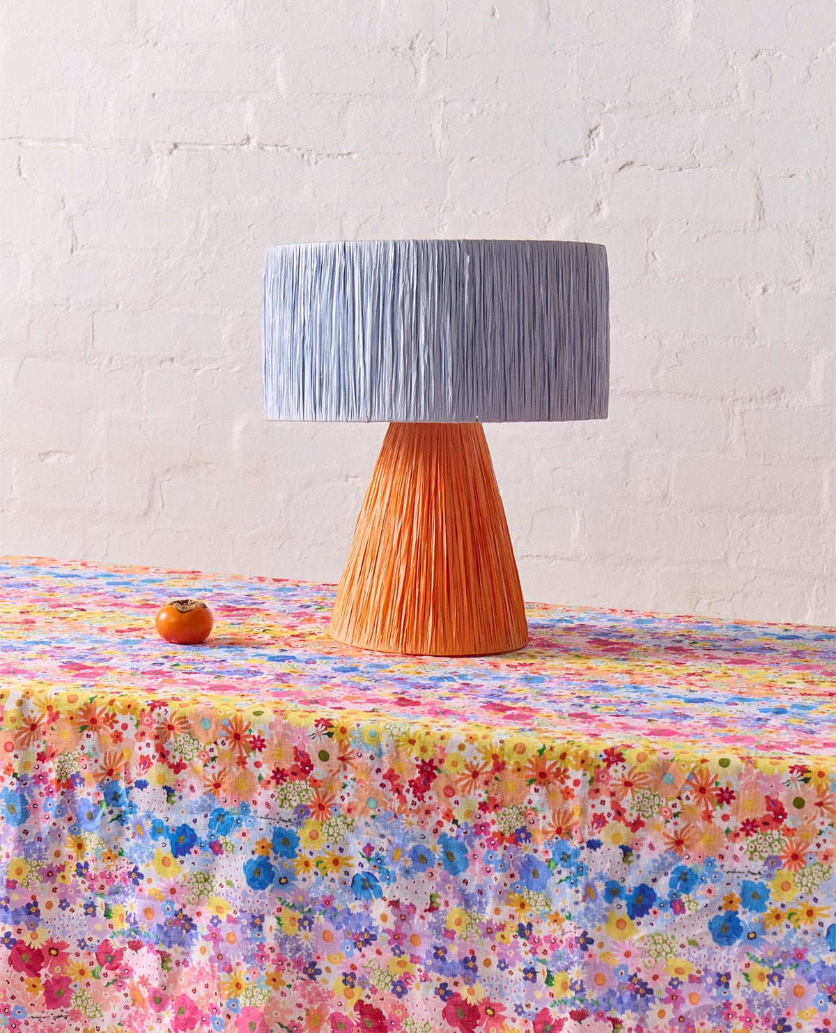 A table lamp with a textured light blue-gray pleated drum shade and a pleated orange conical base, set on a colorful floral tablecloth against a white brick wall.