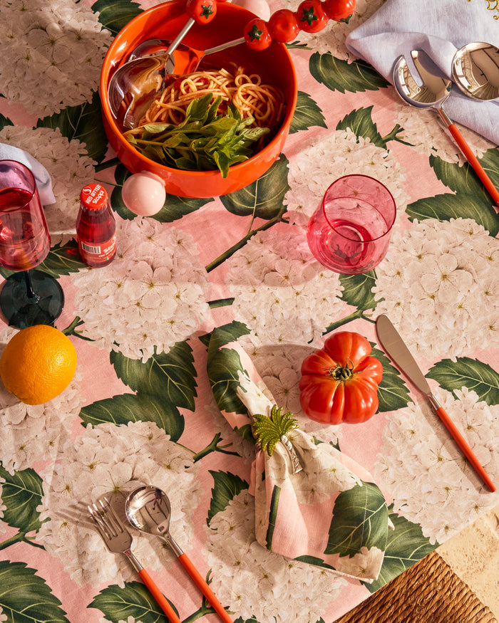 A styled dining table setting featuring a pink and green botanical tablecloth with a matching napkin, pink tinted glassware, silver cutlery with orange handles, and an orange bowl of pasta with greens.