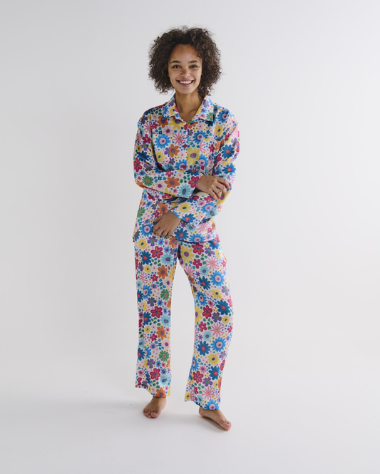 A woman modeling a two-piece pajama set with a colorful, dense floral pattern featuring blue, red, yellow, green, and pink flowers on a white background, photographed against a seamless light gray studio backdrop.