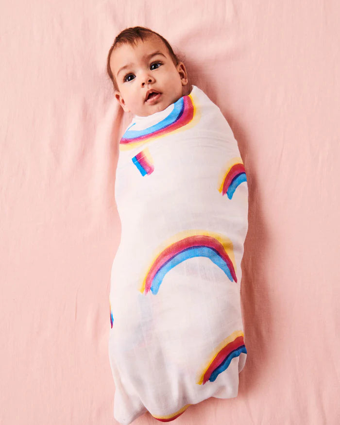 Baby wrapped in a white swaddle with rainbow patterns on a pink background