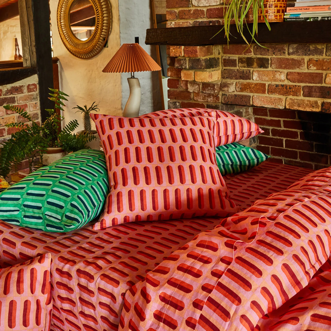 Patterned cushions on a bed against a brick wall with a lamp and plants.