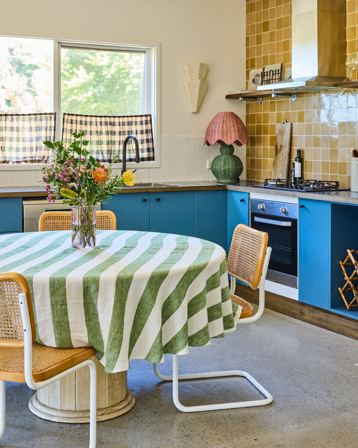 A cozy kitchen interior with bright blue cabinets, a round dining table covered in a green and white striped tablecloth, and woven-back chairs. A window, tiled backsplash, stove, and decorative lamp are visible in the background.