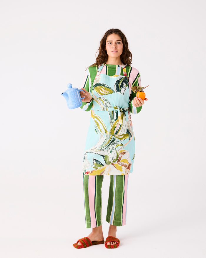 Woman wearing a colorful apron with a floral design, holding a teapot and an orange.