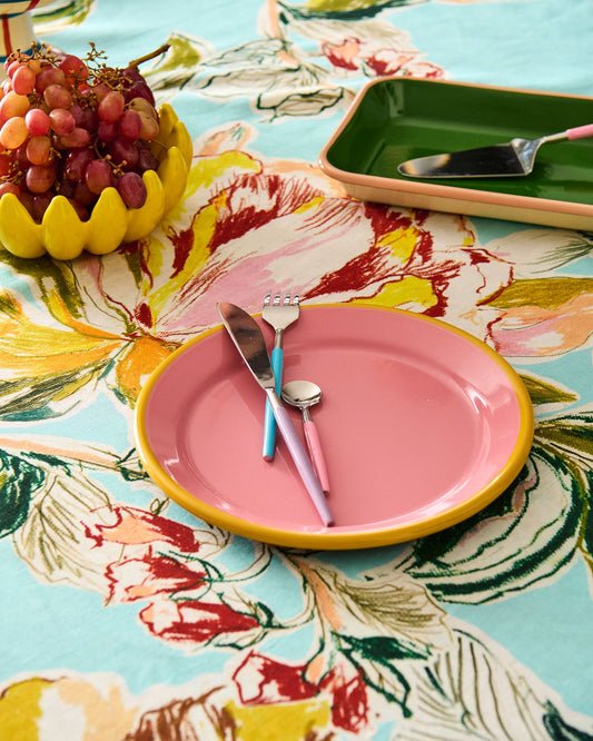 A styled tabletop scene with a pink ceramic plate featuring a yellow rim, set with pastel blue and pink cutlery on a light blue floral tablecloth. A yellow pedestal holds red grapes, and a green tray with a spoon sits in the background.