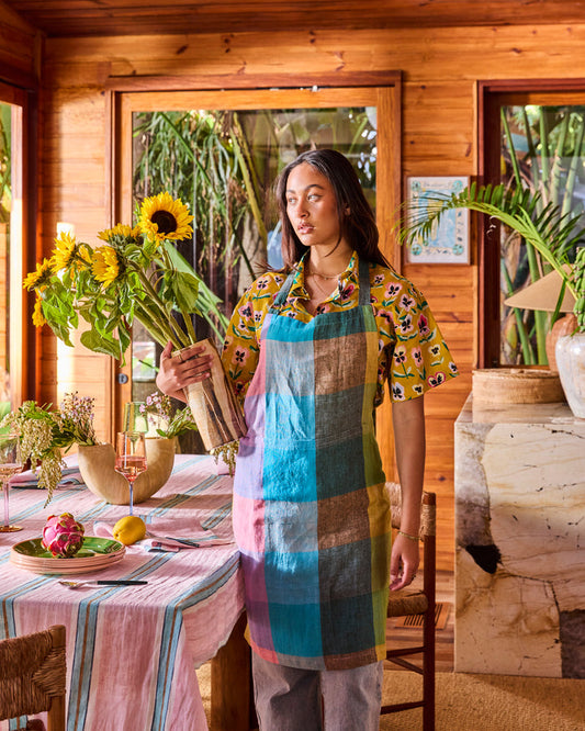 A woman stands in a rustic wooden room holding a large bouquet of sunflowers. She is wearing a colorful plaid apron over a yellow patterned shirt and gray pants.