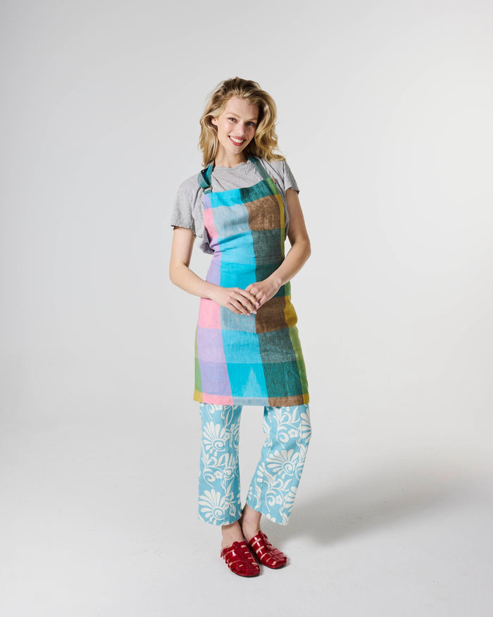 A woman modeling a colorful patchwork apron with a gray t-shirt, light blue floral pants, and red shoes, standing against a light gray studio background.