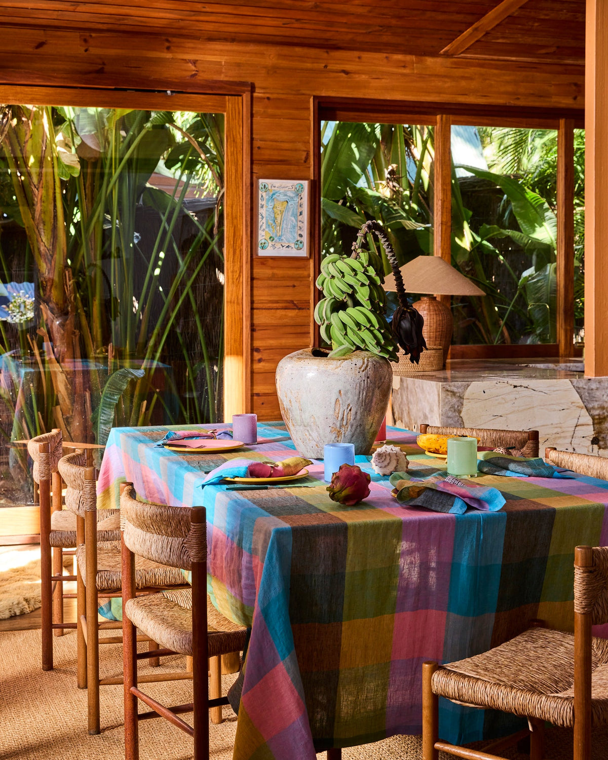 A colorful striped tablecloth in shades of blue, pink, yellow, and green is draped over a dining table set with woven chairs inside a rustic wooden room with large windows overlooking tropical greenery.