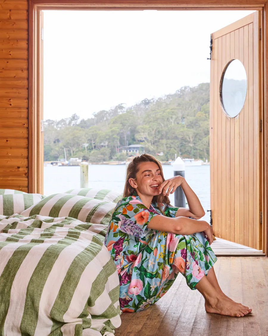 Woman sitting on a bed with striped bedding, looking out of a wooden door to a scenic view.