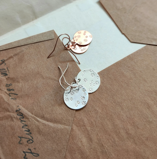Pair of gold and silver earrings with flower stamps on circle disks and earwires, on a textured brown surface.