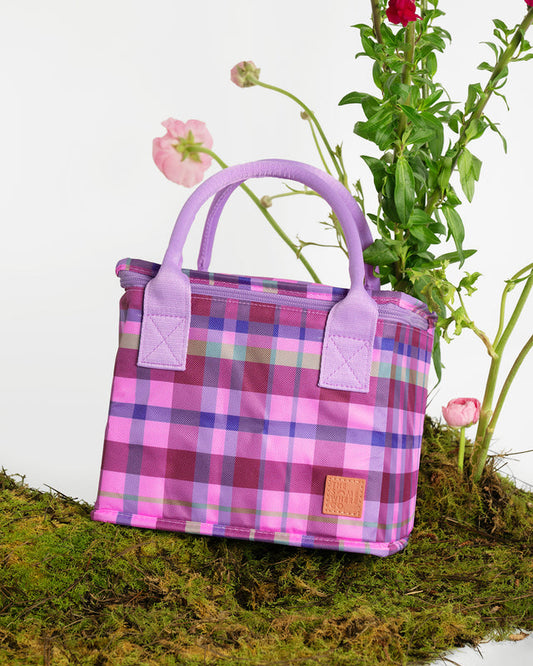 A pink and purple plaid insulated tote bag with lavender handles, styled on moss with flowers against a white background.