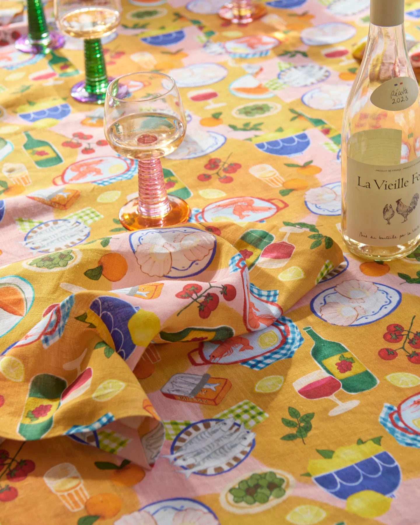 Supper print tablecloth laid out with matching napkins draped on top, scattered wine glasses and wine bottle.