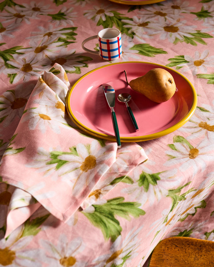 A styled tabletop scene with a pink plate, silver cutlery, a pear, and a striped mug placed on a pink floral tablecloth with matching napkins.