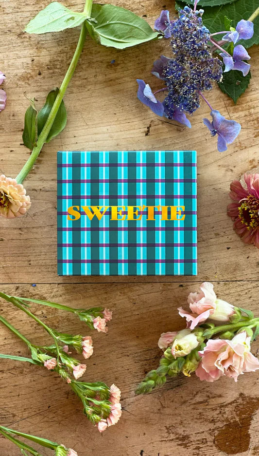 Boxed soap bar with green and blue plaid packaging, gold foil detail on front of box "sweetie". Flowers sit around box on timber.