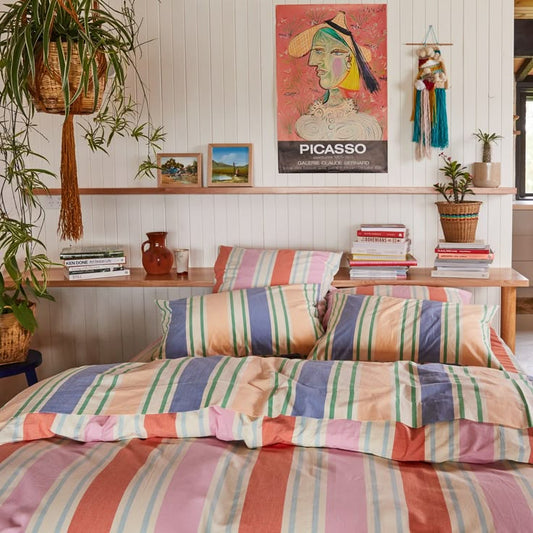 Stripe quilt cover, sheets and pillowcases matching colours on bed with white timber feature wall behind and exposed timber shelves filled with stacks of books and leaning artwork.
