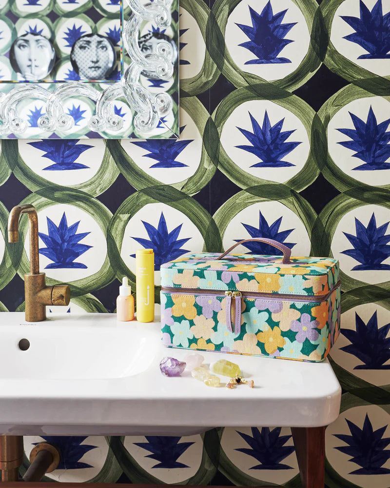 Toiletry case sits on white bathroom vanity. Green base of case with yellow, lilac and blue flowers pattern. Lilac leather handle and straps to open zip. Blue and green patterned wallpaper on wall.