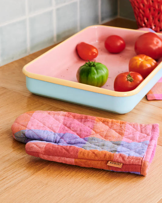 Colourful oven mitt in rainbow tartan print, colourful oven tray filled with tomatoes sit on top timber benchtop with small blue square tiles in background.