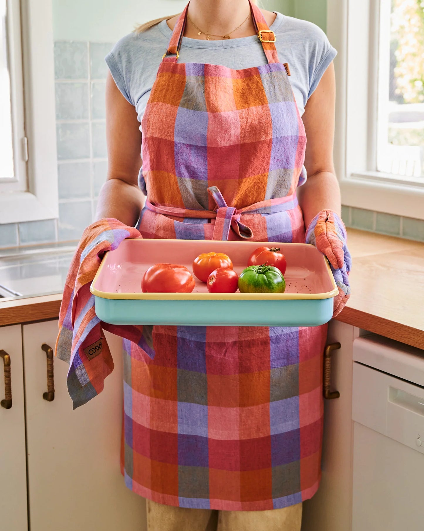 Woman stands wearing tutti frutti rainbow tartan apron, holding oven tray with matching oven mitt and tea towel. Stands in front of bench top and blue tile wall. N