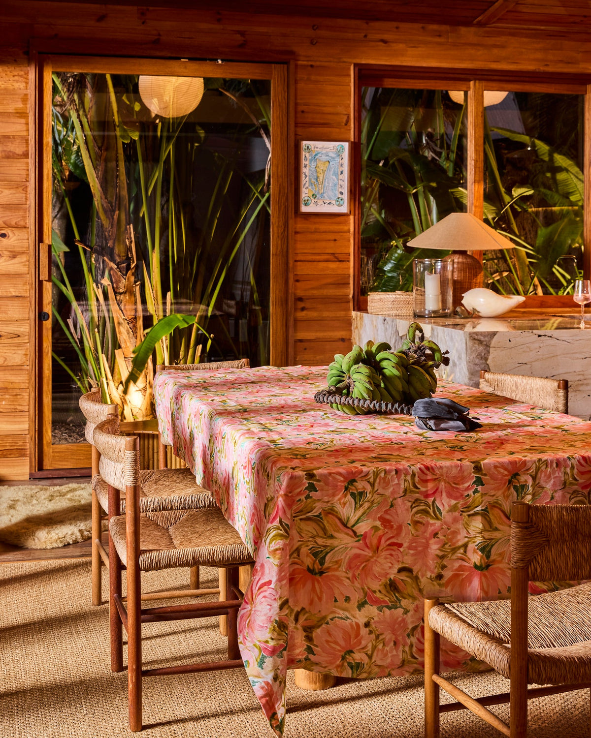 A dining table covered with a pink tropical floral tablecloth, surrounded by woven chairs in a rustic wooden interior with large windows overlooking lush greenery.