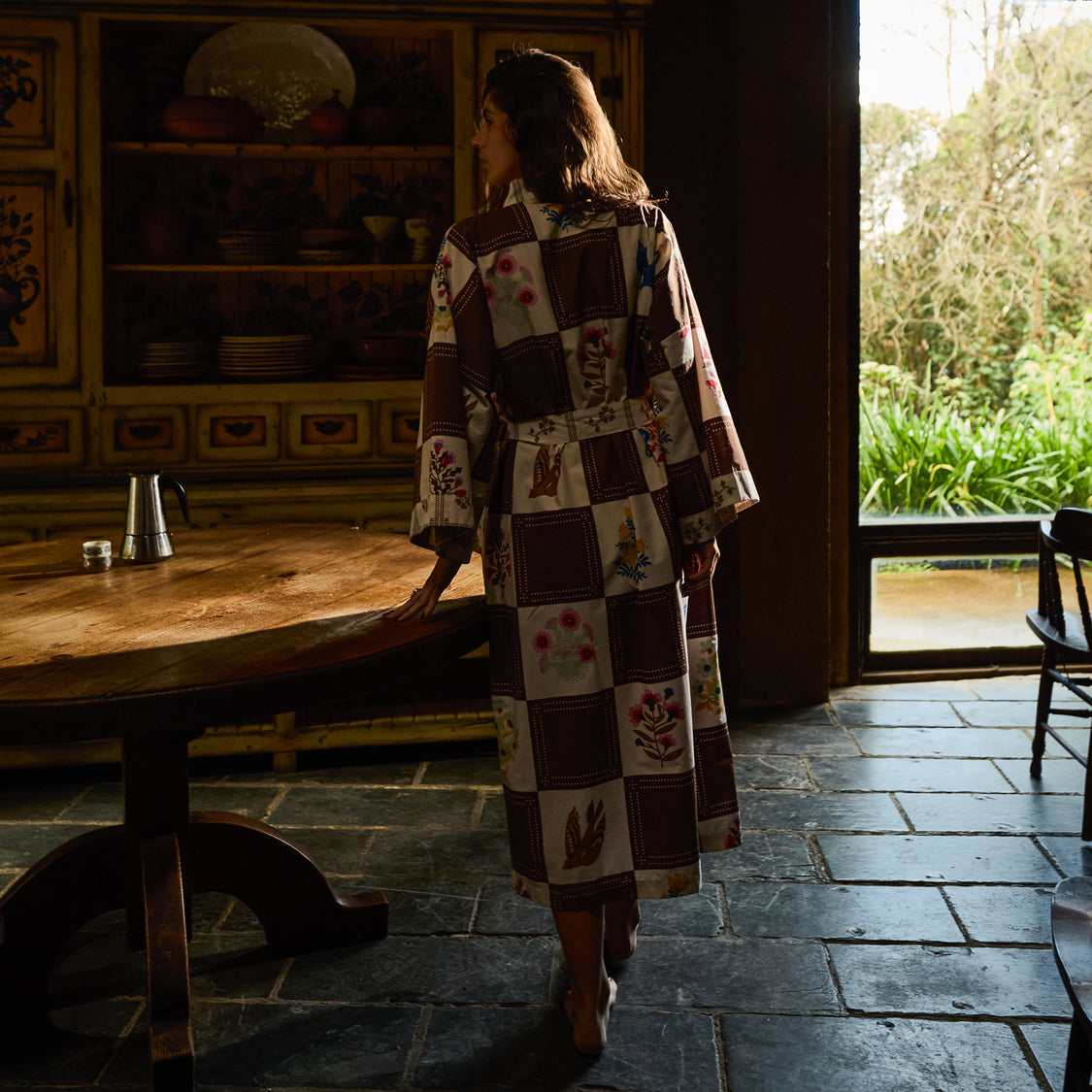 A woman wearing a long patchwork-patterned robe stands barefoot in a rustic room with dark stone floors, a wooden table, and a large window looking out onto greenery.