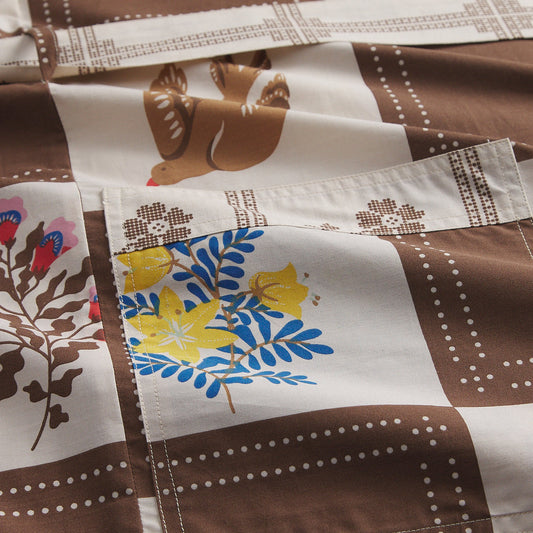 A close-up of a patchwork quilt with brown and cream sections, featuring a brown bird, floral motifs, and a pocket with yellow flowers and blue leaves.