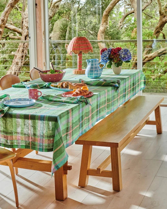 Green tartan tablecloth on timber table with blue ceramic jug, matching napkins blue plates, pink salad bowl and vase sit on top of table scattered. Timber bench seats sits along length of table and big glass window doors in background highlighting green tree views.