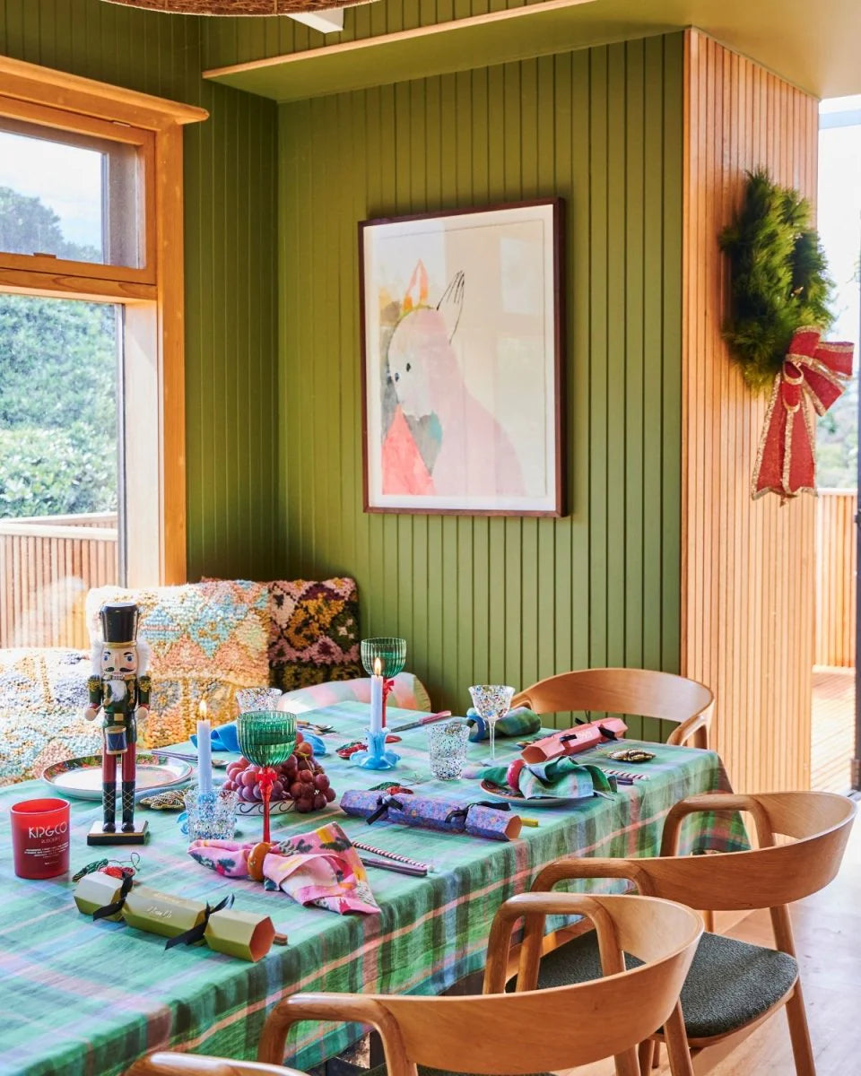 Tartan tablecloth styled on table with assorted glassware, candle, plates, matching napkins. Green featured wall with artwork in background, bench seat filled with Kip and Co cushions and green christmas wreath on joining wall.
