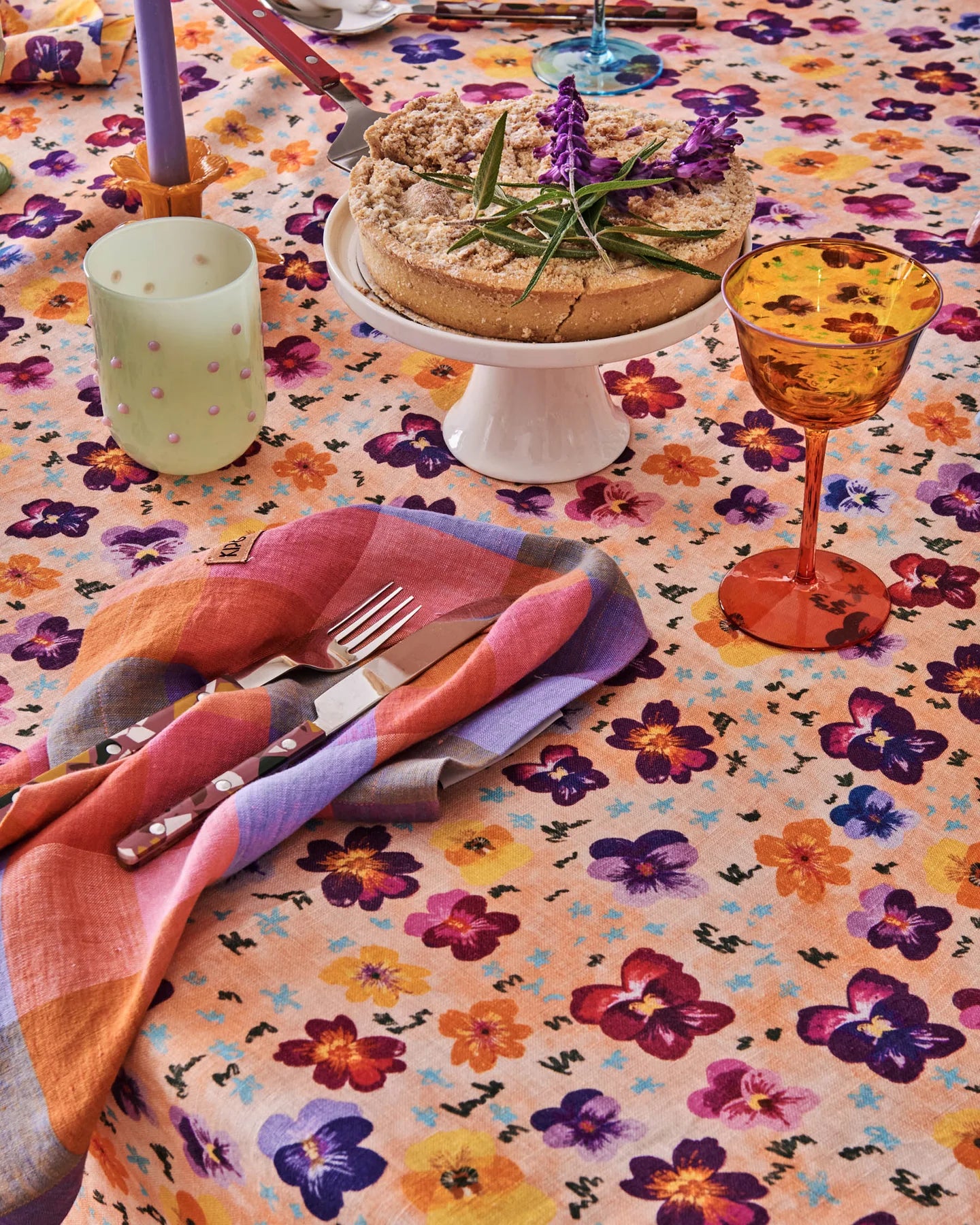 Pansy floral tablecloth with cake stand and glassware sat next to rainbow napkin, sat on top of napkin is cutlery set.