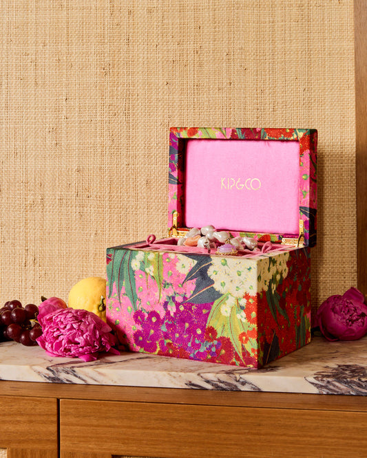 A colorful floral-patterned jewelry box by KIP&CO is open on a marble-topped surface, with pink jewelry inside and decorative flowers and fruit around it against a woven beige backdrop.