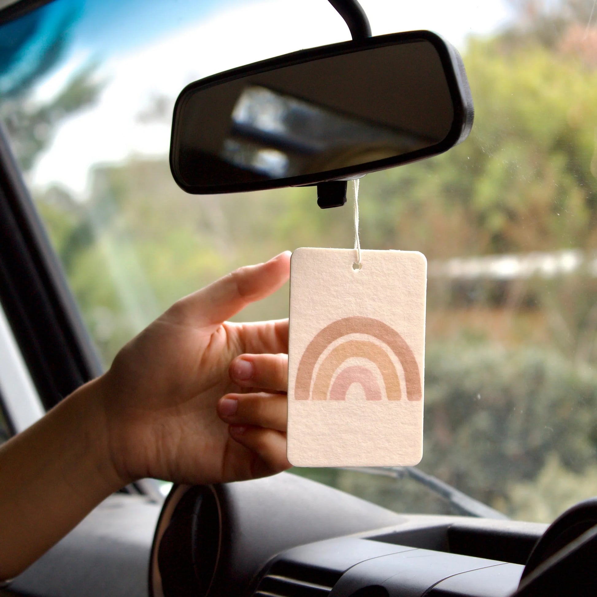 Hand holding a rainbow-patterned car air freshener near a car mirror.