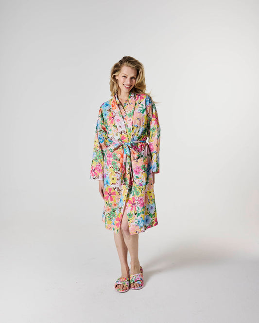 A woman modeling a long-sleeved, tie-front robe with a vibrant multicolor floral print, paired with matching floral slide sandals, against a plain light gray studio background.