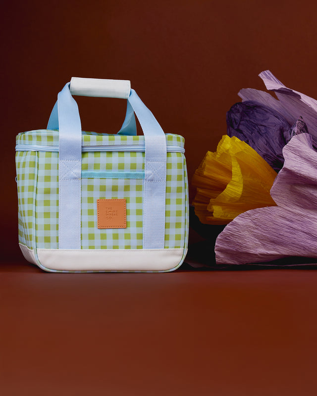 A square canvas lunch bag with a green and white gingham pattern, light blue handles, and a white bottom base, styled next to a bouquet of purple and yellow paper flowers against a dark brown background.